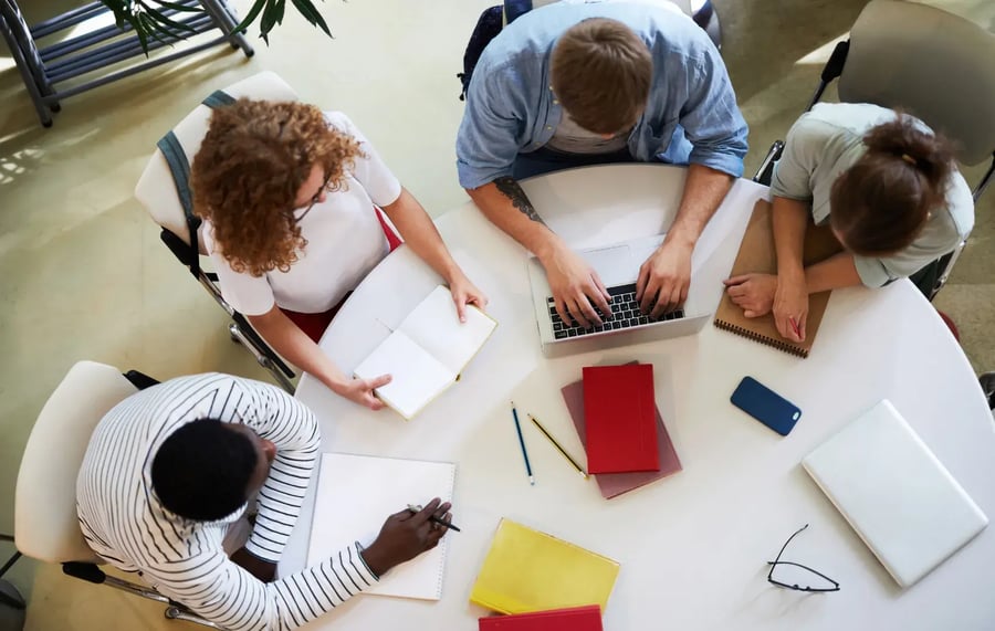 group of students sitting at a round table with their laptops and notes working on school work